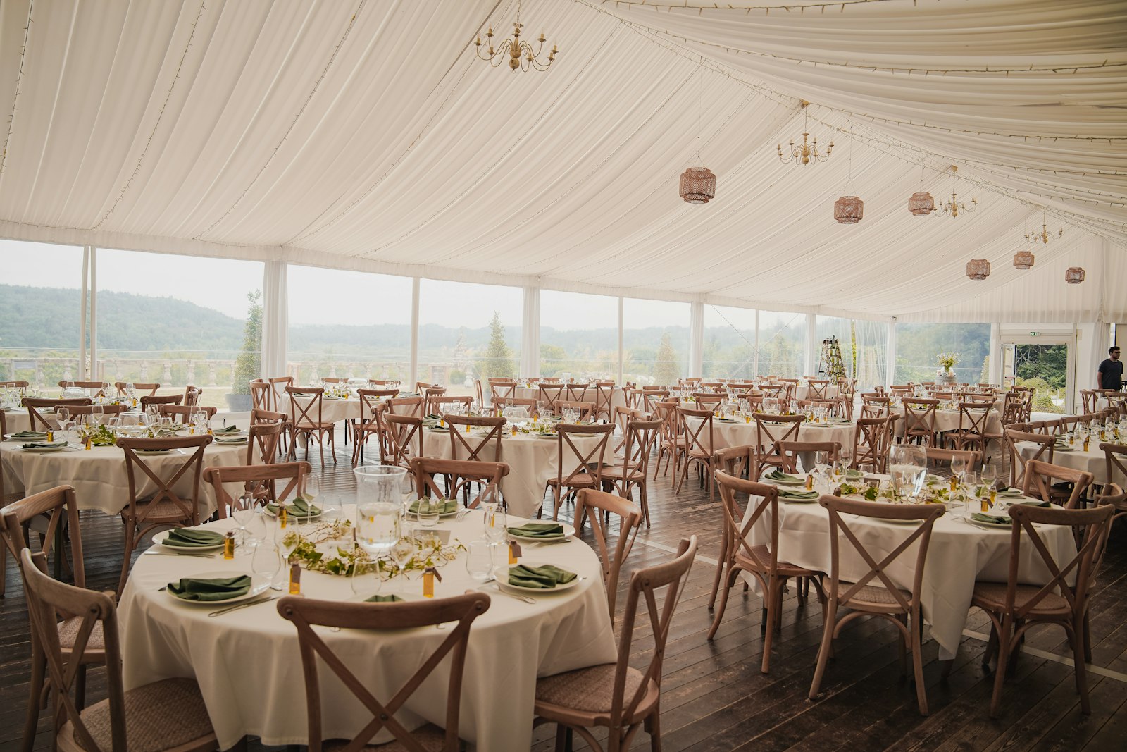 Tented wedding reception set with round tables, cross-back chairs, and greenery centerpieces